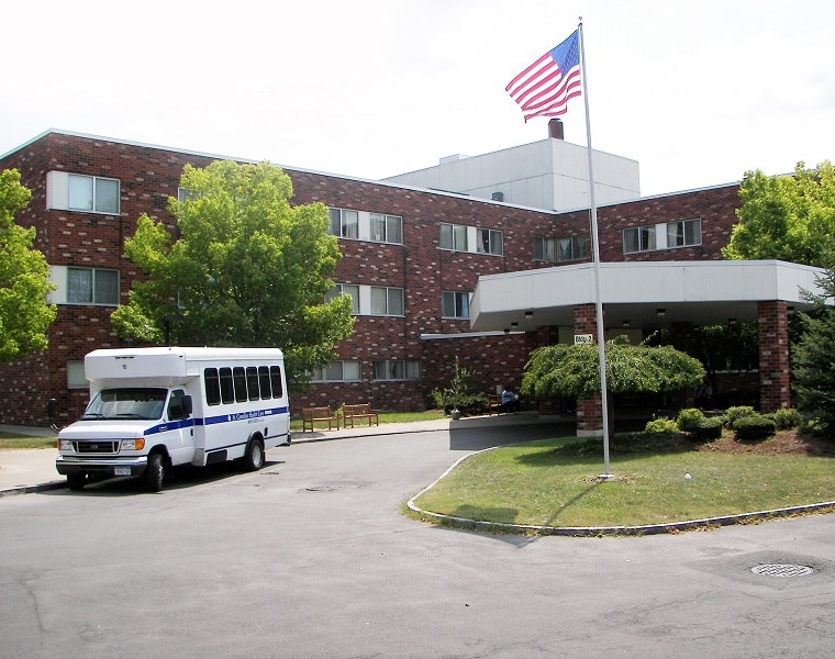 Photo: A brick building with multiple windows, a flagpole, and a shuttle bus parked in front, surrounded by greenery.